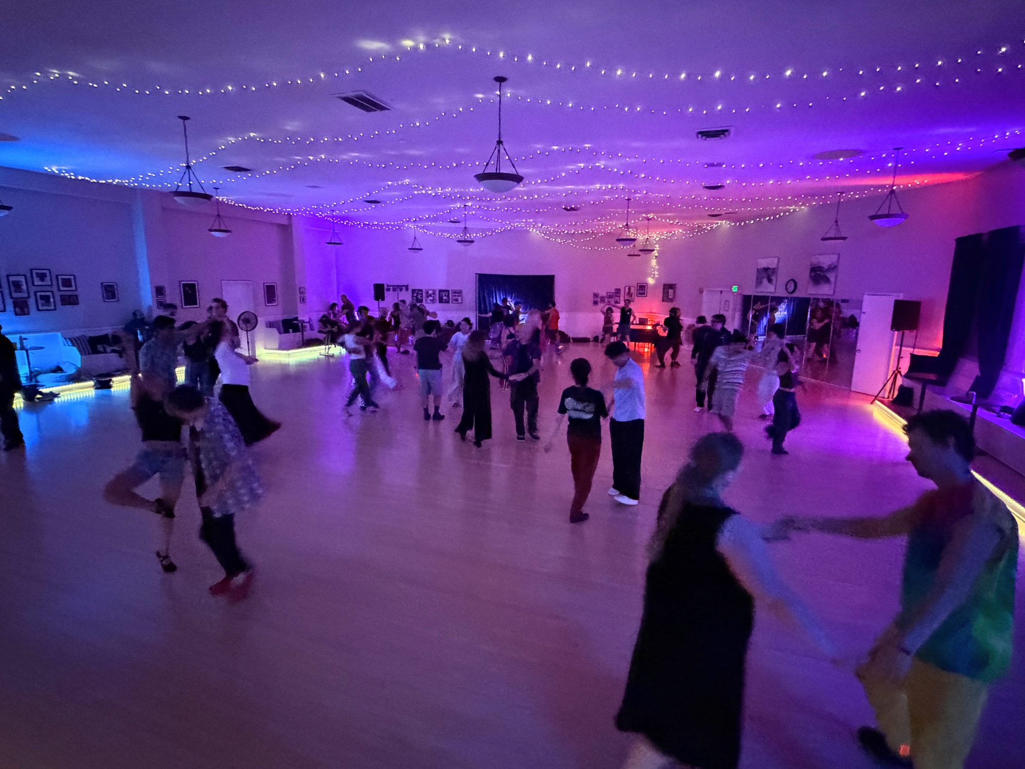 Wide view of the ballroom during a dance: violet and blue light, fairy lights across the ceiling, dancers across the wood floor